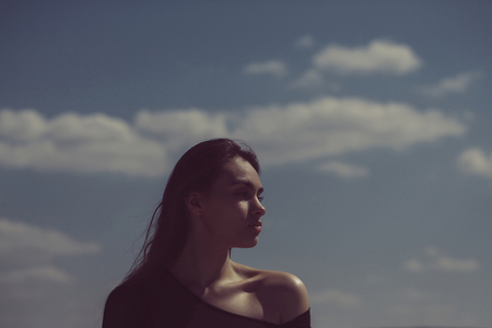 woman or cute girl with long, brunette hair, fashionable model posing in black top with naked shoulder on sunny, summer day on idyllic, blue sky. Creative ideas. Infinity, copy spaceの写真素材