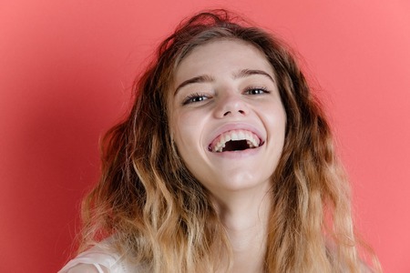 Happy, adorable girl or cute woman with young face, pure skin, no makeup, pretty smile, white, healthy teeth and blond, long, wavy, hair, hairstyle smiling on red wall. Natural beauty and stomatologyの写真素材