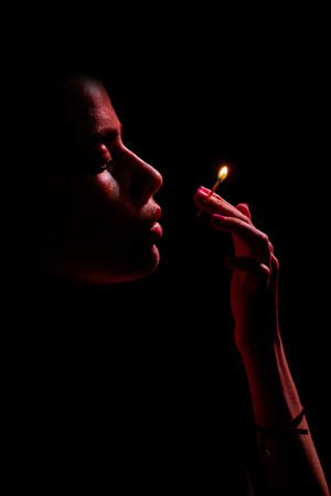 face of adorable girl or pretty woman with oily or wet skin looking at burning match in hand in dark on black background. Light. Idea. Dangerの写真素材