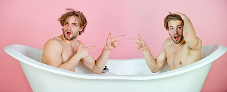 men or unshaven surprised twin brothers with stylish hair, naked, sexy, muscular torso pointing fingers at each other in white bath tub on pink background. Family and love. Spa and hygieneの写真素材