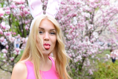 Spring, easter holidays celebration, Happy woman or pretty girl showing tongue, funny face grimace, in bunny ears on blond, long hair at trees with blossoming flowers in park on floral environment.の写真素材