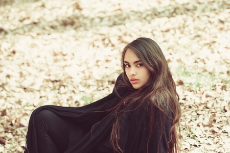 woman. girl or cute woman with long, brunette hair, young face, no makeup, in black coat sitting on ground on sunny day outdoors on natural background. Leisure or vacationの写真素材