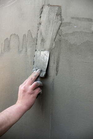 Repairing and finishing work. hand with metallic spatula plastering layer or coat of plaster, stucco, on cement wall surface texture on grey background. Working tool. Construction and renovationの写真素材