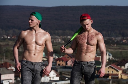 twins, athletic men or handsome athletes with naked, sexy, muscular torsos, six pack, ab, in red and green cap, jeans standing with baseball bat on mountain landscape. Sport and training. Fit, fitnessの写真素材