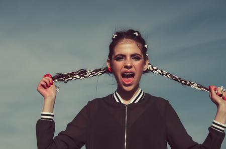 Amazed girl or cute woman shouting with dental braces on teeth in open mouth, stylish makeup and long braids, brunette hair, in hands on grey sky. Stomatology. Beauty and hairdressing salonの写真素材