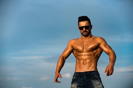 sexy man with muscular wet body and strong torso of bearded bodybuilder athlete in summer sunglasses and jeans posing with bare chest and belly sunny outdoor on blue sky background, copy spaceの写真素材