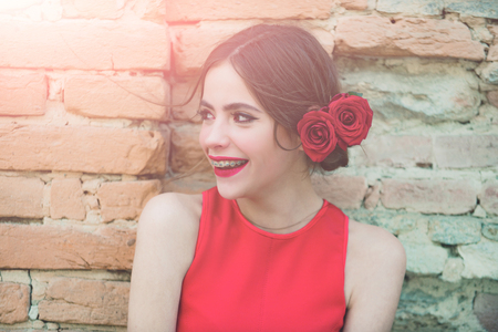 Happy girl or cute woman smiling with dental braces on teeth, red lips, stylish makeup and roses in brunette hair on sunny day outdoors on old brick wall. Stomatology, health. Beauty salonの写真素材