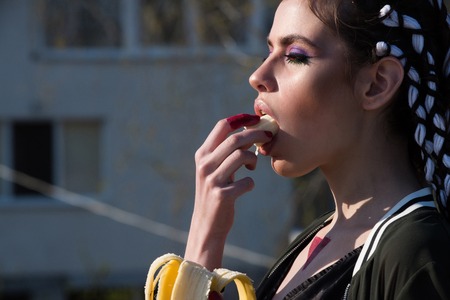 woman with stylish makeup, long braids, brunette hair, eating organic banana on sunny day outdoors on urban environment. Health and vegetarian diet, healthy dietingの写真素材