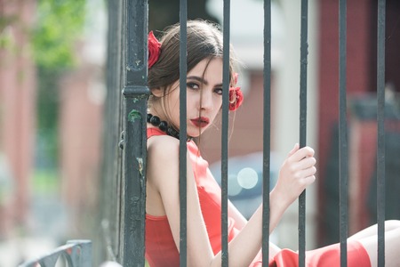girl or spanish woman with fashionable makeup and red rose flower in hair, girl in black beads accessory on iron fence background, beauty and fashionの写真素材