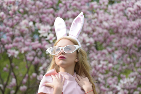 Pretty girl or cute woman, fashionable model, with bunny ears and funny glasses with long, blond hair posing in pink top at blossoming trees in park on blurred, floral environment. Easter. Springの写真素材