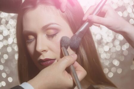 Hands of visagistes putting powder on fashion model face skin with two makeup brushes. Pretty girl or cute woman with closed eyes and stylish eyeshadows, lips on glowing background. Visage, cosmeticsの写真素材