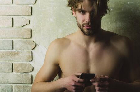 guy or bearded macho with sexy naked chest holding smartphone or mobile phone in muscular hands with biceps, triceps on brick wall. Technology, internet surfing. Fitness, sportの写真素材