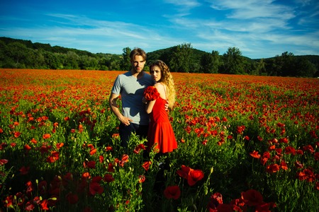 woman and guy with long curly hair in red dress hold flower bouquet in field of poppy seed with green stem on sunny natural background, summer, drug and love intoxication, opium, couple in loveの写真素材