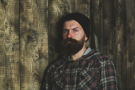 Bearded man or brutal caucasian hipster with long beard and moustache with serious face posing in stylish black hat and plaid jacket on grey wooden wall. Fashion, beauty and lifestyleの写真素材