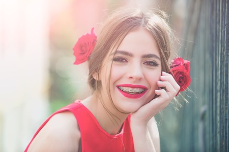 girl or cute woman smiling with dental braces on teeth, red lips, stylish makeup and roses in brunette hair on sunny day outdoor, Stomatology health. Beauty salonの写真素材