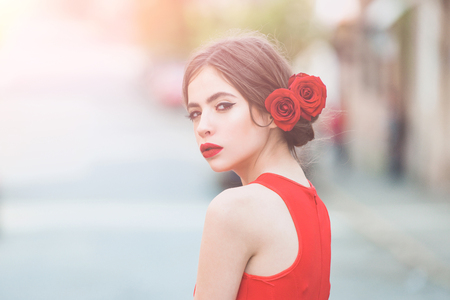 girl or pretty woman, with red lips, makeup on adorable young face and fresh roses flowers in brunette hair posing outdoors on blurred urban environment. Visage. Beauty and hairdressing salonの写真素材