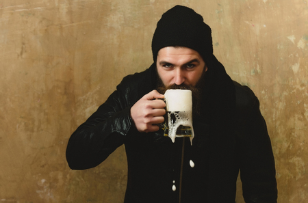 man or bearded hipster with long beard and moustache drinking beer with foam from glass mug in black hat and jacket on beige wall. Alcohol, bad habits, addictive, lifestyle, refresher, conviveの写真素材