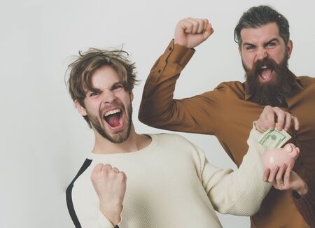 men with beards or friends laughing and putting money dollars in piggy bank with winner gestures on grey background. Savings, budget, finance, cash and moneyboxの写真素材