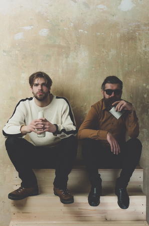 man with milk or yogurt bottle and bearded model in sunglasses with stainless hip flask. Two friends in casual wear sitting with drinks on wooden stairs. Healthy lifestyle. Friendship, conviveの写真素材