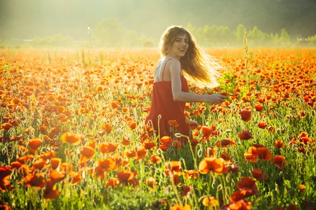 pretty woman or happy smiling girl with long curly hair in red dress in flower field of poppy seed with green stem on natural background, summer, spring, drug and love intoxication, opiumの写真素材