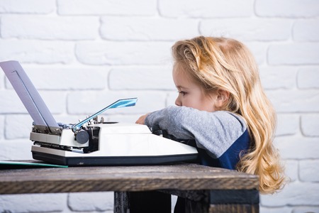 writer kid or small boy or businessman child with blonde hair sitting at table and typing typewriter with paper on white brick wall background, business and new technologyの写真素材