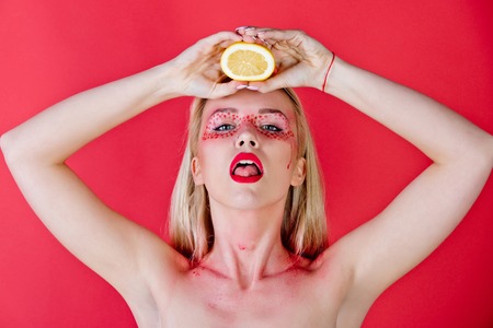 girl with creative fashionable makeup on face hold and drink lemon fruit in hands on red background, beauty and fashion, allergy, healthcare, vitamin, vegetarian and dietingの写真素材