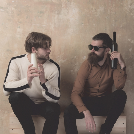 friendship of stylish friends talking and drinking on beige wall. Handsome model in sweater with milk or yogurt bottle and bearded man in sunglasses with stainless hip flask. Healthy lifestyleの写真素材