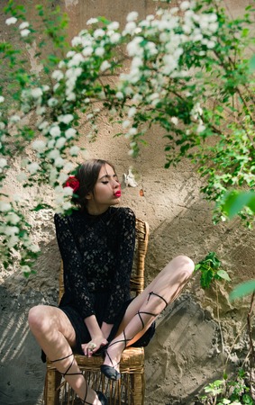 girl or fashionable young model with red lips, roses in hair in black, lace dress sitting on wicker chair under bush with white, blossoming flowers on beige outdoor wall. Springの写真素材