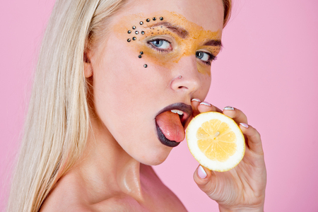 woman with creative fashionable makeup on face hold and lick lemon fruit in hands on pink background, beauty and fashion, allergy, healthcare, vitamin, vegetarian and dietingの写真素材
