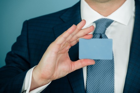 hand holding blank bank or business card in formal suit with stylish tie on blue background. Economy, credit, ecash, banking, ethics and informationの写真素材