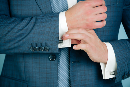 Male hands fixing stylish silver cufflinks on white shirt cuffs sleeves in formal suit with elegant buttons on blue background. Business fashion and jewelryの写真素材