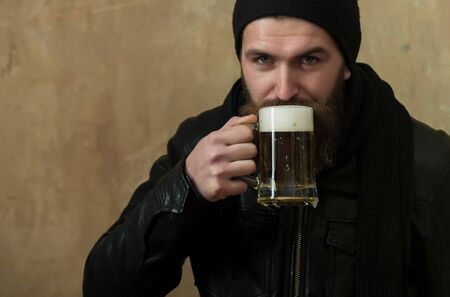 hipster with long beard and moustache drinking beer with foam from glass mug in black hat and jacket on beige wall. Alcohol, bad habits, addictive, lifestyle, refresher, conviveの写真素材