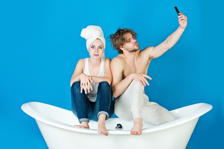 woman and man with towel sitting on white bathtub on blue background make selfie photo with mobile phone, family and relationship, spa and beauty, relax and hygiene, blogging, couple in loveの写真素材