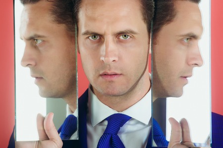 Man with three faces in two mirrors. Reflections of handsome businessman or manager with blue tie on red background. Agile business. Male beautyの写真素材