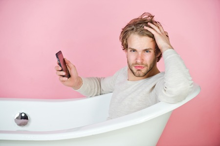 handsome young man in underwear sitting in white bathtub with mobile phone on pink background, spa...