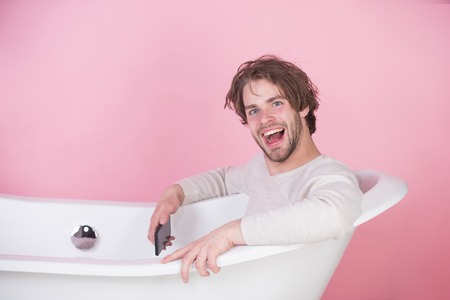 man smiling in underwear sitting in white bathtub with mobile phone on pink background, spa and beauty, relax and hygiene, healthcare, new technology and social networkの写真素材
