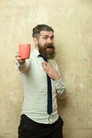 manager or bearded man with long beard and stylish hair on happy face in tie and white shirt on textured beige background hold tea or coffee cup, morning and energy, refreshmentの写真素材
