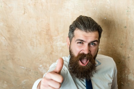 bearded man or hipster with long beard and stylish hair on happy face in tie and white shirt on textured beige background with thumb up gesture, copy spaceの写真素材