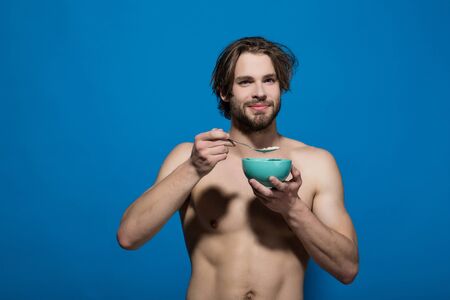 healthy food and dieting, man with bare chest eating breakfast of oatmeal with milk in bowl with spoon on blue background, fitness, morning, copy spaceの写真素材