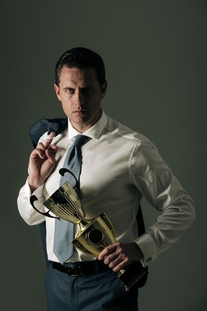 champion man, successful businessman or manager holding golden winner cup in fashion white shirt, tie and jacket on shoulder on grey background. Business and success. Victory and trophyの写真素材