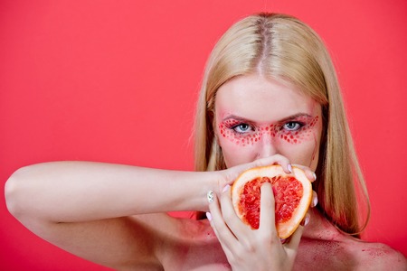 grapefruit in hands of pretty blonde woman with creative fashionable makeup on face hold fruit on red background, beauty and fashion, allergy, healthcare and vitamin, vegetarian and dietingの写真素材