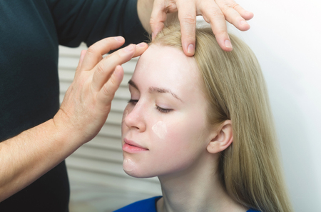 Visagiste hands applying concealer on woman face skin with closed eyes. Cute girl or fashionable model with long blond hair in beauty salon. Visage, makeup, cosmetics and skincareの写真素材