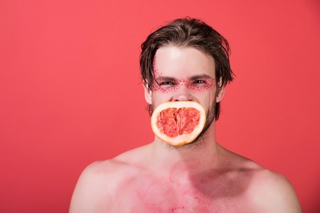 man with creative fashionable makeup on face hold grapefruit fruit in mouth on red background, beauty and fashion, allergy, healthcare and vitamin, vegetarian and dietingの写真素材