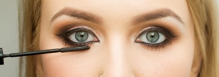 Girl with eye makeup. Brush applying black mascara on eyelashes. Woman face with healthy young skin on grey background. Beauty salon. Visage and make up. Cosmetics and skincareの写真素材