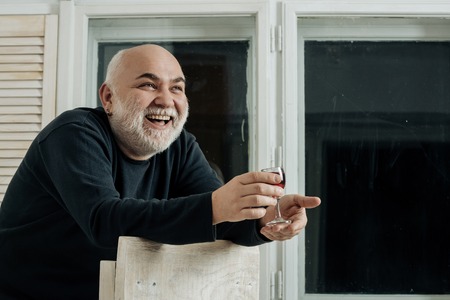 Happy man smiling with glass of wine. Senior person or pensioner with grey beard in black sweater in room on window background. Alcohol and appetizer. Unhealthy lifestyle and bad habitsの写真素材
