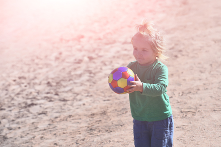 child or small boy with cute smiling face and long blond hair playing with colorful ball on background of sand beach in sunny day. Happy childhood, copy spaceの写真素材