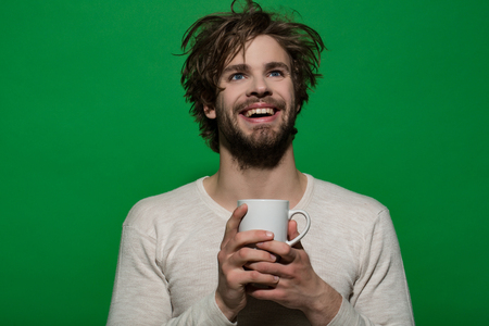 smiling man with cup of tea or coffee has uncombed hair in underwear on green background, morning refreshment and drinkの写真素材