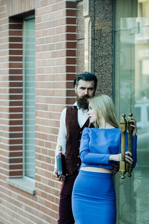 Boyfriend with bottle hugging girlfriend holding glass door. Cute girl in blue dress and hipster with wine outdoors on brick wall. Alcohol and convive. Couple in love. Unhealthy lifestyle. Bad habitsの写真素材