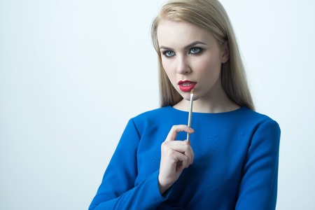 Girl with makeup brush at red lips. Sexy woman with blond long hair wearing blue dress isolated on white. Fashionable model with bright make up on face skin. Fashion. Visage and cosmetics, copy spaceの写真素材