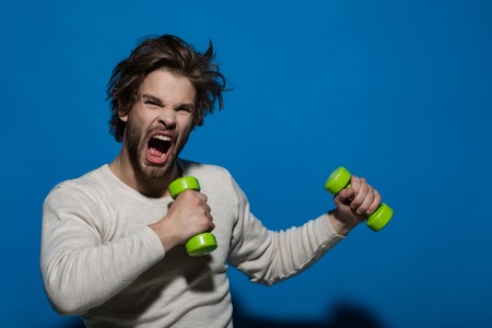 shouting man with barbell or dumbbell workout, has disheveled and uncombed long hair and beard on face in white underwear on blue background, morning exercise and wake up, barbershop, sportの写真素材